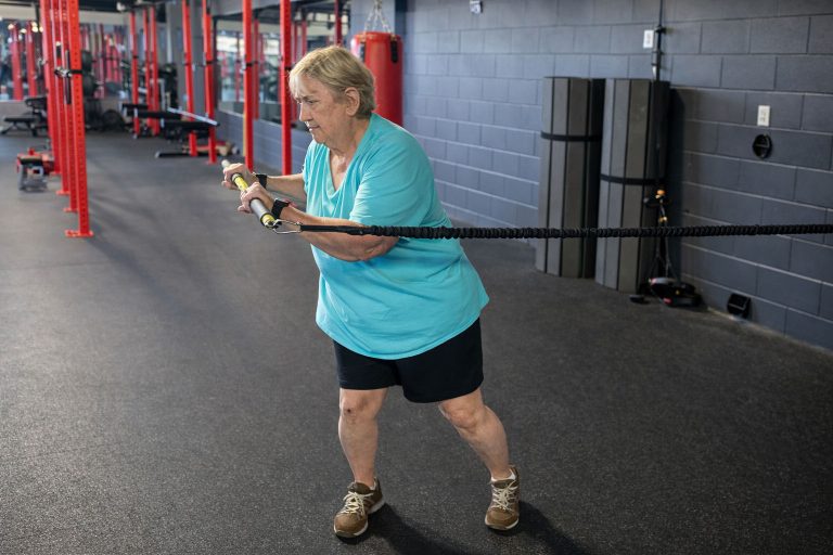 Older female client performing a resistance band row exercise at a personal training gym with red squat racks, demonstrating one-on-one training at Rug'd Training in Mount Pleasant, South Carolina.