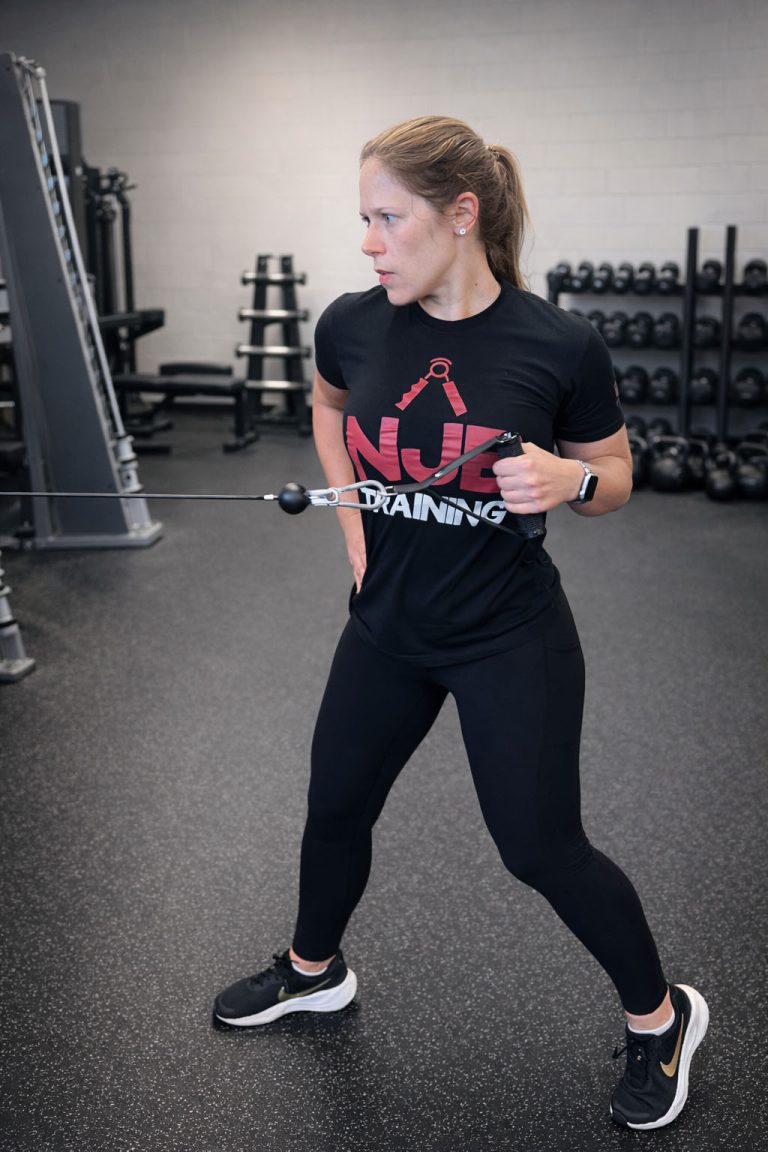 Woman performing a functional training exercise using a resistance band in a personal training gym, with weight racks and equipment in the background at Rug'd Training in Mount Pleasant, South Carolina.